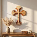 A wooden wall cross with layered organic cutouts, displayed on a beige wall above a wooden console table with pampas grass, books, and a candle.