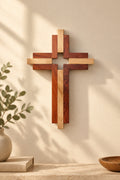 A wooden cross wall decor piece with layered wood strips in natural light brown, reddish-brown, and dark brown, mounted on a solid beige wall.